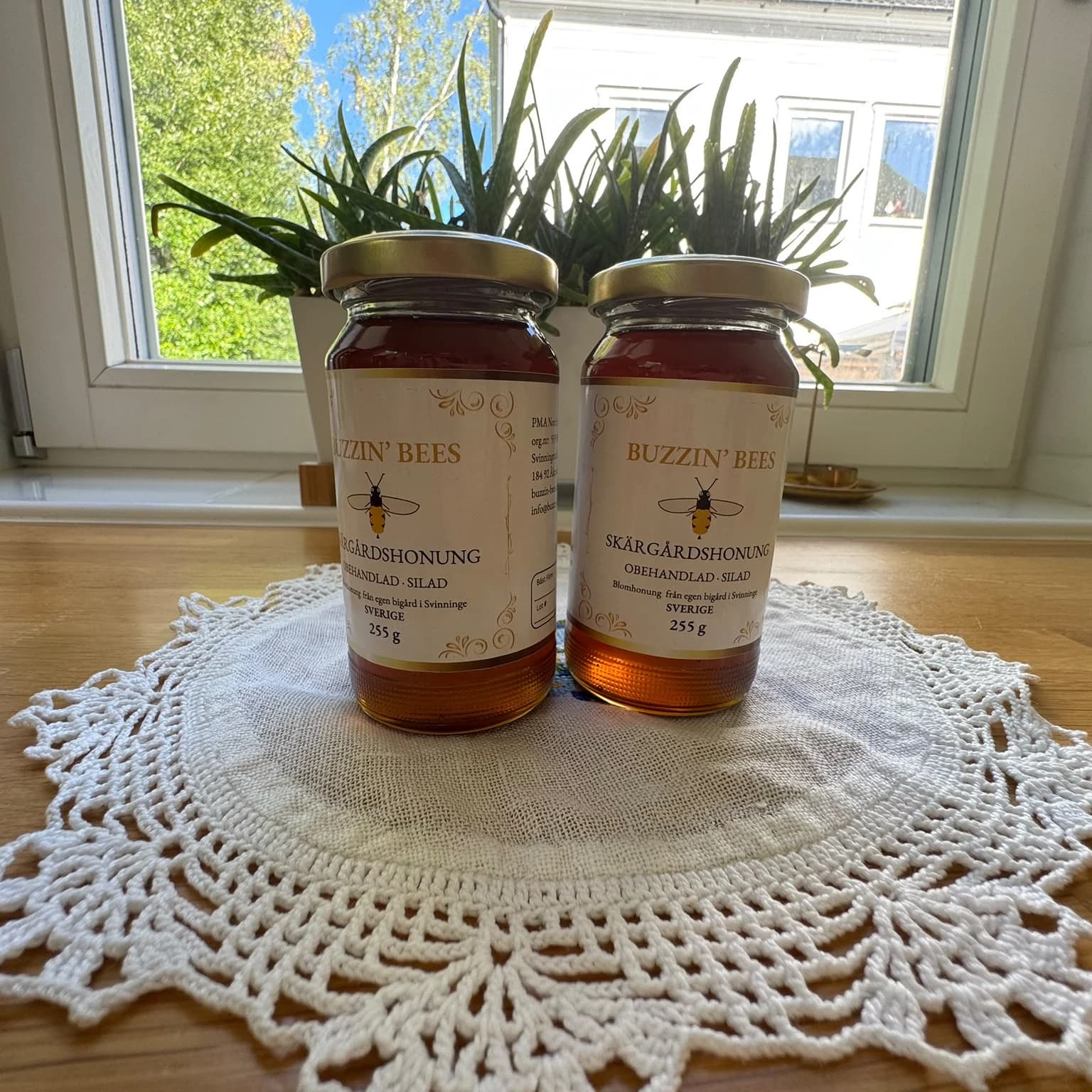 Two jars of Buzzin' Bees Sensommarhonung in front of a sunlit window, on a crocheted cloth, the Skärgårdshonung label visible
