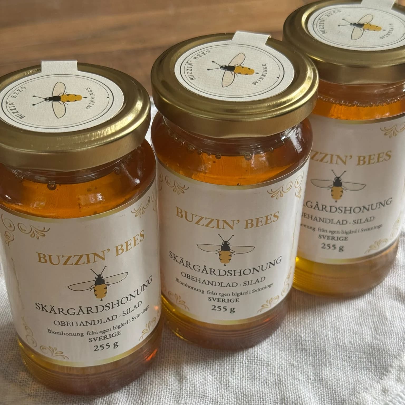 Three jars of Buzzin' Bees Skärgårdshonung seen from above at an angle, lids and labels visible, on a crocheted cloth